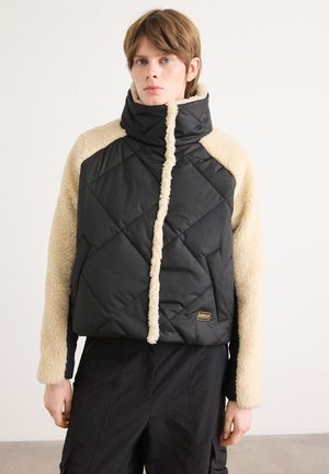 Zwarte gewatteerde pufferjack met beige nep-bonten mouwen en kraag, met contrasterende texturen en een klein logo-label aan de voorkant.