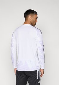 Camisola desportiva de mangas compridas em branco com padrões geométricos subtis, com riscas pretas nos ombros e gola redonda.