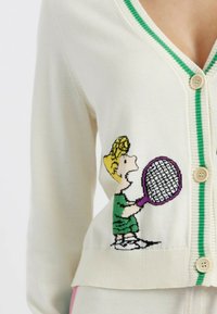 Crème kleurige cardigan met groene afwerking, met een geborduurd kind dat een tennisracket vasthoudt. Knopen van lichtgekleurde kunststof.