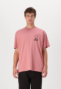 Camiseta de algodón rosa con mangas cortas, cuello redondo y un diseño gráfico negro en el lado izquierdo del pecho que presenta un árbol y texto.