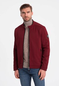 Burgunderfärgad jacka med ståkrage, framzipper och texturerat tyg. Har ett märke på vänster axel och sidfickor.