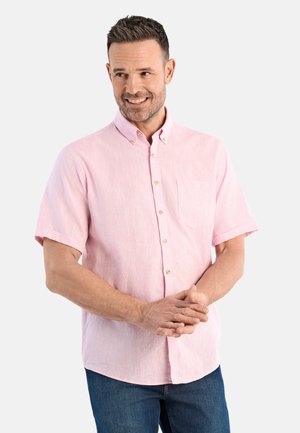 Lachende man met kort haar, gekleed in een lichtroze overhemd met korte mouwen en blauwe spijkerbroek, met zijn handen voor zich samengevouwen.