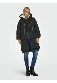 Doudoune noire avec capuche, dotée d'une doublure en fausse fourrure et de fermetures éclair. Assortie à un pull en tricot et à un jean en denim.