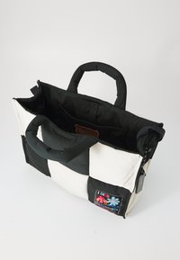 Geantă tote în carouri alb-negru, cu mânere căptușite, un interior spațios și o patch-uri colorate care prezintă flori și text pe o latură.