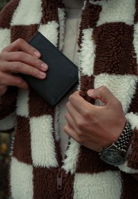 Black leather wallet held in hands, wearing a brown and white checkered fleece jacket with a textured surface and a zipper detail.