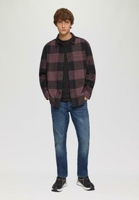 Chemise à carreaux en violet foncé et noir, superposée à un t-shirt noir. Un jean en denim bleu et des baskets noires complètent la tenue.