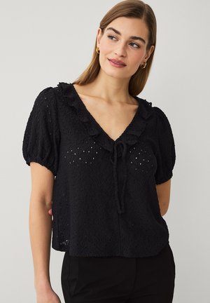 Frau mit glattem hellbraunem Haar trägt eine schwarze, strukturierte Kurzarmbluse mit Rüschen am V-Ausschnitt und Bindendetail, kombiniert mit schwarzen Hosen.