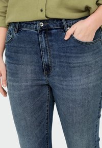 Jeans de mezclilla azul oscuro con un acabado desgastado, que presentan un cierre de cremallera y un botón. Corte regular con bolsillos frontales y detalles sutiles de costura.