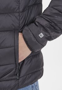 Veste noire rembourrée avec texture matelassée, poche zippée et poignets élastiques. Présente un petit logo argenté sur la manche.