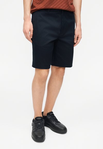 Homme portant un short bleu marine foncé jusqu'aux genoux, des baskets noires et une chemise texturée couleur rouille, les bras détendus le long du corps.