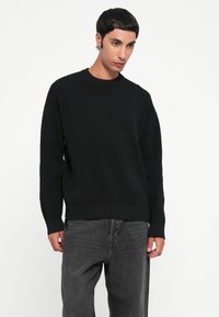 Svart ribbestrikket genser med rund hals, senkede skuldre og en strukturert overflate, sammen med mørk grå denimbukser.