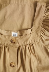 Blusa di cotone color beige con scollatura quadrata, dettaglio plissettato e due bottoni marroni sul davanti. Etichetta visibile con il marchio "GAP".