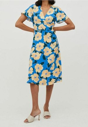 Femme portant une robe midi bleue à grandes fleurs jaunes et manches courtes, associée à des sandales à talons ouvertes beige.