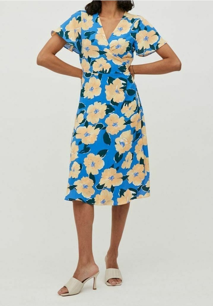 Femme portant une robe midi bleue à grandes fleurs jaunes et manches courtes, associée à des sandales à talons ouvertes beige.