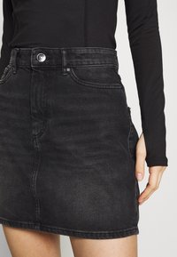 Czarna spódnica dżinsowa z wysokim stanem, zapinana na guziki z przodu, z bocznymi kieszeniami. Zniszczona faktura z widocznymi detalami szycia.