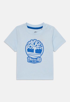 Jasnoniebieska koszulka z krótkim rękawem wykonana z bawełny, z niebieskim logo drzewa i tekstem "Timberland Est. 1973" na środku. Zapięcie na zatrzask przy ramieniu.