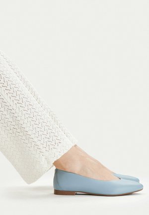 Pies con zapatos planos azul claro combinados con pantalones blancos texturizados sobre un fondo blanco.