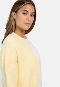 Geel gebreide cardigan met een ontspannen open ontwerp, zachte textuur en vleermuismouwen, gedragen over een witte top.