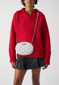Maglione rosso a maglia con colletto, abbinato a una gonna plissettata nera a quadri. Porta una borsa a tracolla bianca trapuntata con una catena.