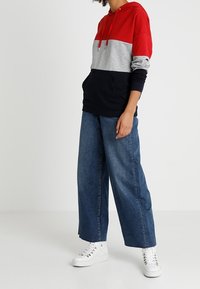 Felpa con cappuccio a blocchi di colore rosso, grigio e blu navy con tasca frontale; abbinata a jeans di denim blu a gamba larga e scarpe da ginnastica bianche.