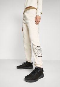 White sweatpants with ribbed waistband and black DKNY logo detail on the left leg, paired with textured black athletic shoes.