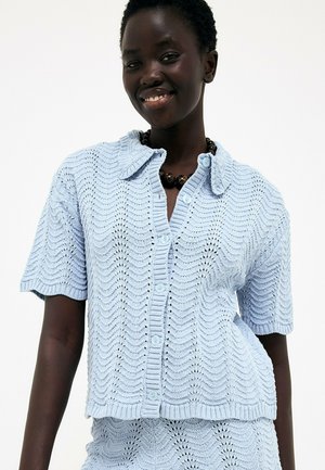 Frau trägt eine hellblaue, gestrickte, kurzärmelige, geknöpfte Strickjacke mit Wellenmuster und eine Perlenkette und lächelt in die Kamera.