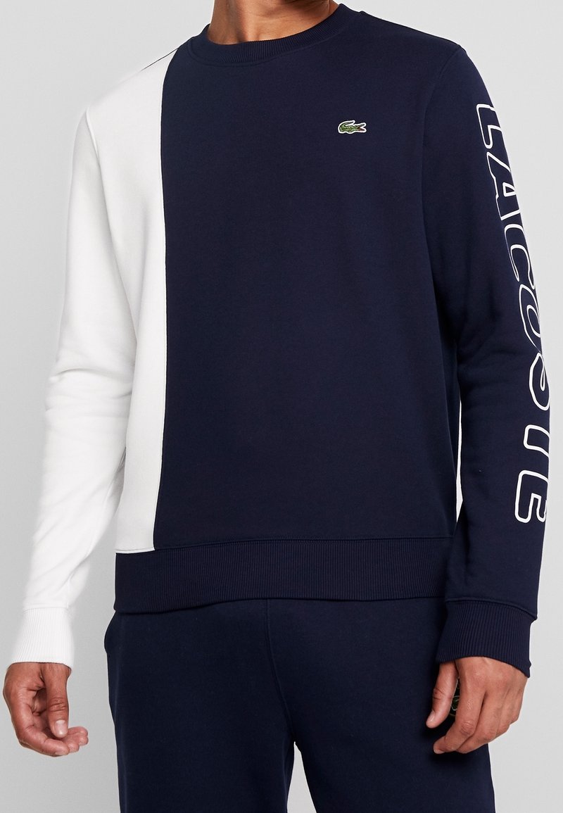 Felpa color-block in blu navy e bianco con polsini e orlo a coste, caratterizzata da un piccolo logo verde e dalla scritta "LACOSTE" stampata su una manica.