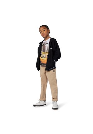 Veste zippée noire, t-shirt graphique avec scène urbaine, pantalon beige et baskets blanches avec accents noirs. Tenue simple et décontractée.