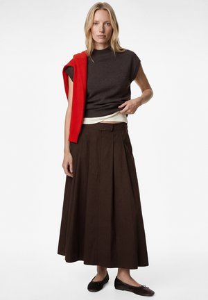 Frau trägt ein dunkelbraunes ärmelloses Oberteil, langen plissierten braunen Rock, schwarze Ballerinas und hat einen roten Pullover über eine Schulter gelegt.