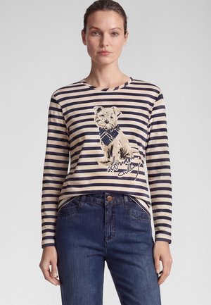 Frau trägt ein langärmeliges cremefarbenes und marineblau gestreiftes Shirt mit einem Hunde-Motiv und dunkelblaue Jeans vor einfachem Hintergrund.