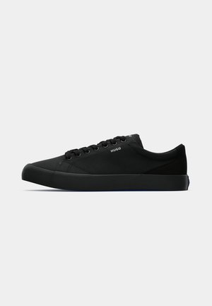 Des baskets noires en toile présentent une texture lisse, une pointe arrondie, un design à lacets avec six œillets, et une épaisse semelle en caoutchouc noire avec des accents bleus.