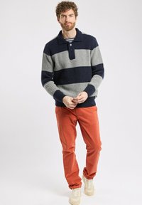 Pull en maille rayé bleu marine et gris, doté d'une texture côtelée, d'un col et d'une patte de boutonnage à trois boutons. Associé à un pantalon orange et des baskets.