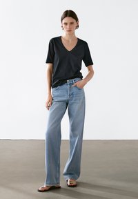 Kvinde iført sort V-udskåret T-shirt, lyseblå brede bukser og sorte sandaler mod en ensfarvet hvid baggrund.
