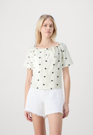 Lys grøn cropped top med sort blomsterbroderi, der har en samlet halsudskæring og korte ærmer, parret med hvide denimshorts.
