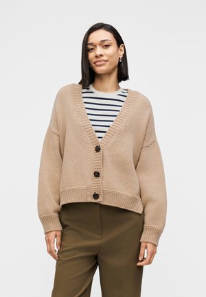 Beige gestrickte Strickjacke mit V-Ausschnitt, ausgestattet mit vier dunklen Knöpfen und gerippten Bündchen. Trägt man über einem gestreiften Hemd und olivgrünen Hosen.