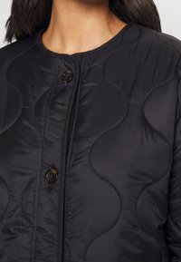 Černá prošívaná bunda s kulatým výstřihem zdobená vlnitým vzorem a dvěma předními knoflíky. Hladká textura s lehkým provedením.