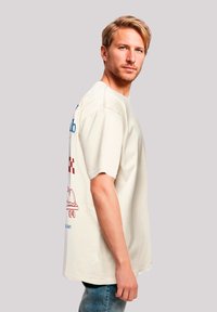 Light beige oversized t-shirt made of cotton, featuring blue and red graphics on the back, short sleeves, and a relaxed fit design.