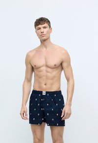 Marineblå boxer shorts med et mønstret design, elastisk talje, og lille logo-patch foran. Let bomuldsstof, løs pasform.