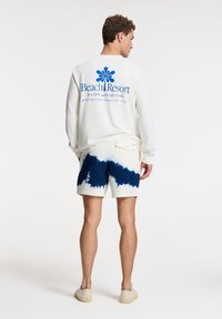 Camisa blanca de mangas largas con gráfico azul de "Beach Resort"; pantalones cortos de baño con diseño de tie-dye azul y blanco; calzado casual.