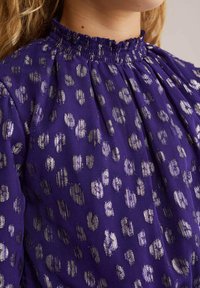 Ljubičasta bluzica sa nabranim izrezom, s teksturiranim zlatnim točkicama kroz cijelu površinu. Tkanina izgleda lagano i blago sjajno.