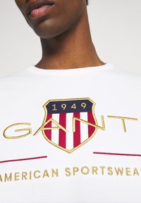 Weiße Sweatshirt mit einem rot-blauen Wappenlogo und goldener Stickerei, Text mit der Aufschrift "GANT" und "AMERICAN SPORTSWEAR", mit runder Ausschnitt.