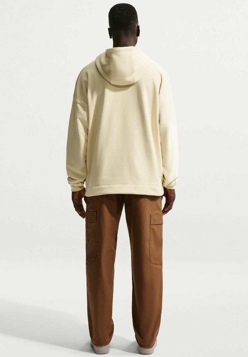 Un homme est debout de dos, portant un hoodie crème et un pantalon cargo marron sur un fond clair uni. Mains détendues le long du corps.