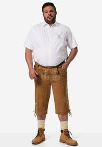 Lederhosen marroni in suede con ricami floreali, abbinate a una camicia bianca a maniche corte e stivali beige con lacci, con calze color crema.
