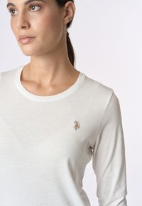 Camicia bianca a maniche lunghe realizzata in tessuto morbido, con scollatura rotonda e un piccolo logo ricamato in oro sul petto sinistro.