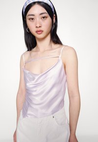 Camisola de satin lilás com decote drapeado, alças finas e design drapeado; usada com calças de cor clara. Textura suave com brilho subtil.