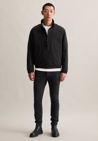 Jonge man staat in een zwarte zip-up jas, witte shirt, zwarte jeans en zwarte laarzen tegen een beige achtergrond.