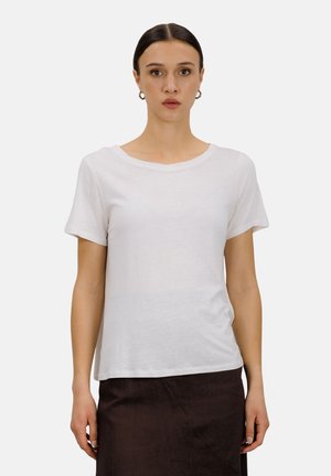 Kvinne med mørkt hår samlet bak, iført sølvfargede runde øreringer, en enkel hvit kortermet skjorte og et mørkt skjørt foran en hvit bakgrunn.