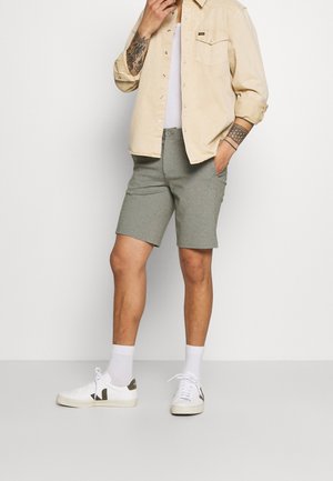 Helle beigefarbene Hemd über einem weißen T-Shirt, kombiniert mit grauen Shorts. Weiße Schuhe mit dunklen Akzenten. Tätowierungen am Arm sichtbar.