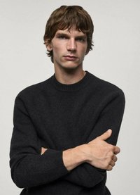 Schwarzer gestrickter Pullover mit rundem Halsausschnitt, strukturiertem Muster und langen Ärmeln, getragen von einer Person mit verschränkten Armen in einer neutralen Umgebung.