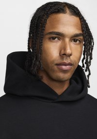 Sort hætte sweatshirt med en blød tekstur, der har en rummelig hætte og kengurulomme, båret af en person med fletninger.